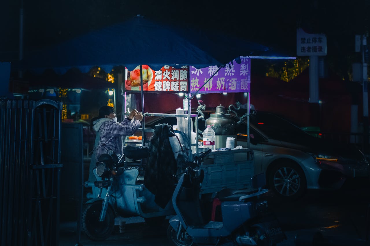 A bustling night market food stall in an urban street with vibrant lighting and a vendor preparing food.